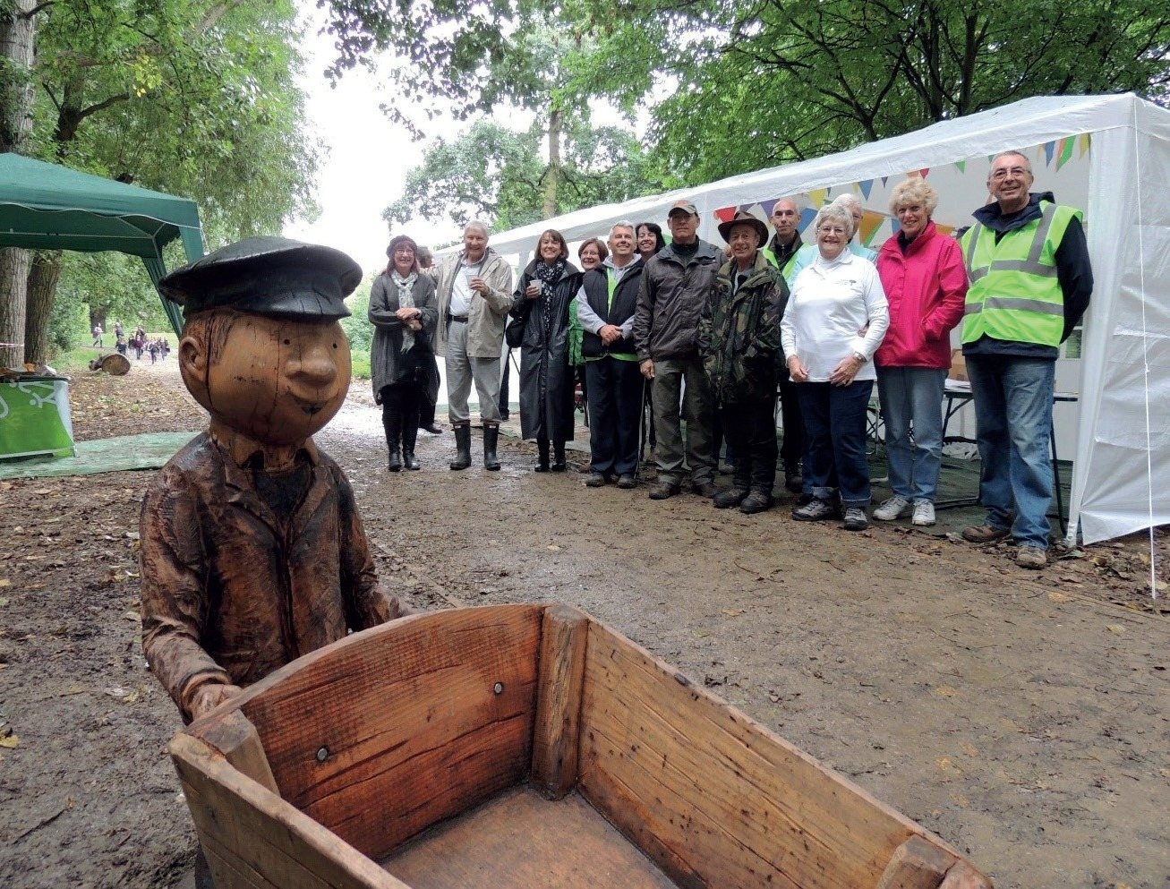 A carved wooden figure with a wheelbarrow, with a group standing in a row behind it
